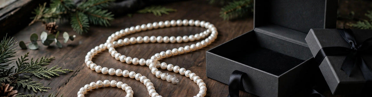 White freshwater pearl necklace and pearl bracelet beside an open dark gift box with evergreen sprigs on a walnut surface, a masculine holiday gift scene